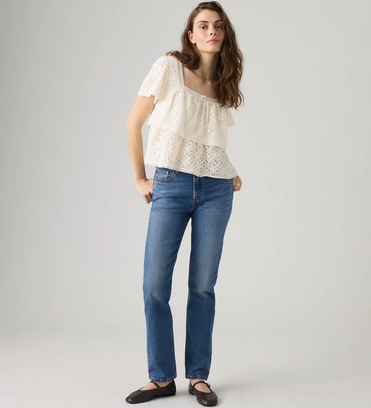 A woman modeling a short-sleeve cream lace top and blue jeans against a light gray studio background.