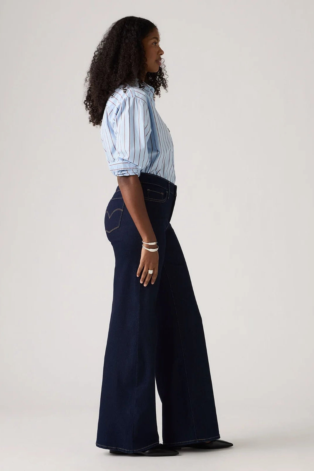 A woman stands in profile wearing a light blue and white vertically striped short-sleeve blouse with puffed shoulders and dark navy wide-leg denim pants, against a plain light gray studio background.